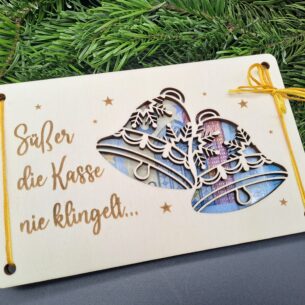 Zweiteilige Holzkarte aus Birkensperrholz mit dem Schriftzug „Süßer die Kasse nie klingelt“ und ausgeschnittenen Glocken-Motiven, durch die Geldscheine sichtbar sind. Die Karte ist mit einer gelben Schnur fixiert und liegt auf Tannengrün.