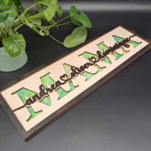 Ein längliches, dreilagiges Holzschild mit dem ausgeschnittenen Wort "MAMA". Dahinter ist ein grün-blau gewolkter Hintergrund sichtbar. Im Vordergrund stehen die verbundenen Kindernamen "andrea, oliver & dominique" in dunkler Ebenholz-Lasur.