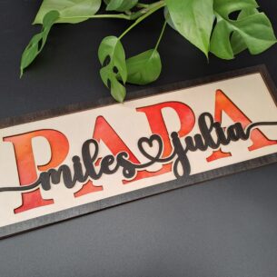 Ein längliches, dreilagiges Holzschild mit dem ausgeschnittenen Wort "PAPA". Dahinter ist ein rot-orange gewolkter Hintergrund sichtbar. Im Vordergrund stehen die verbundenen Kindernamen "miles & julia" in dunkler Ebenholz-Lasur.