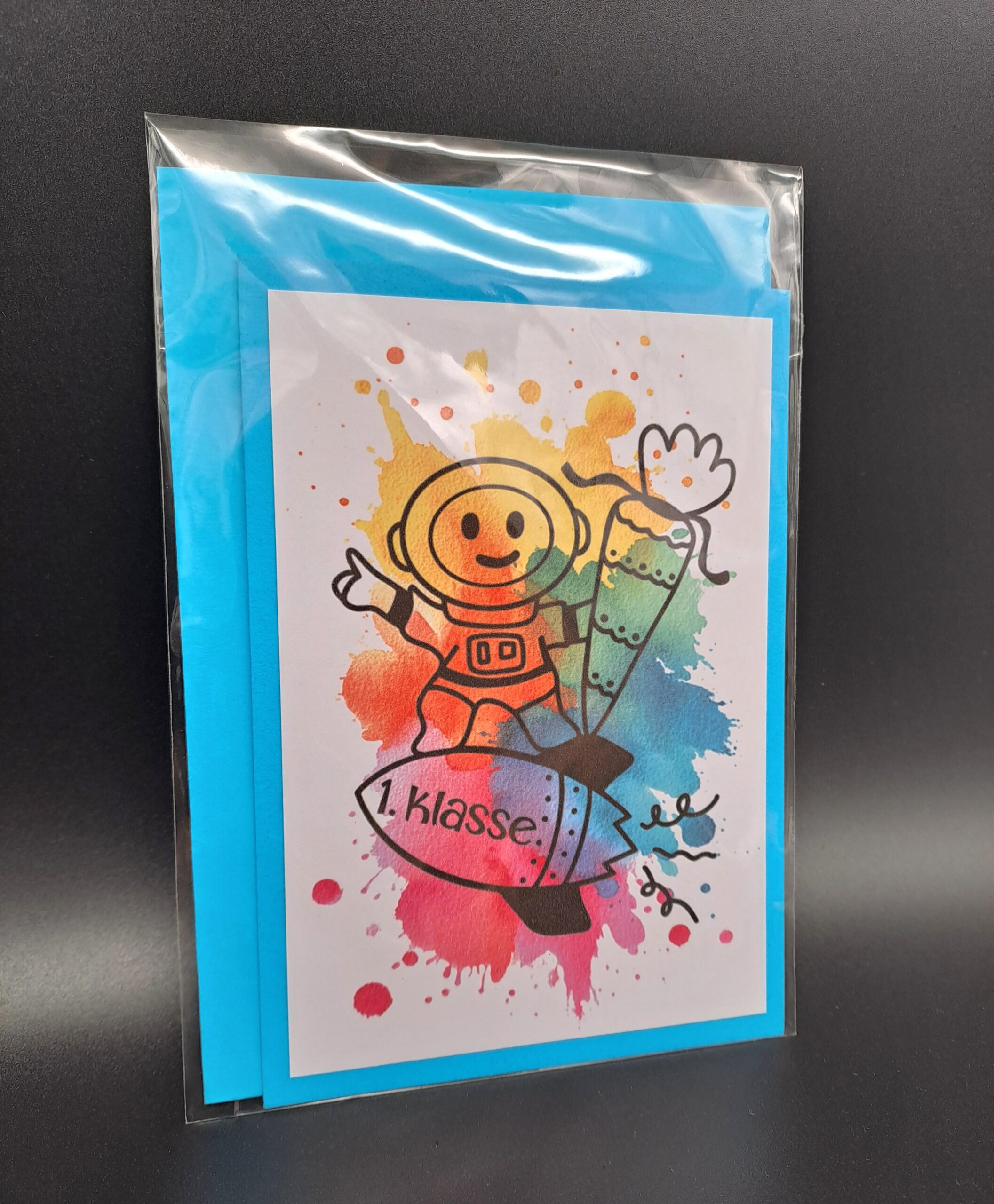 Eine in Cellophan verpackte A6 Klappkarte zur Einschulung mit einem blauen Umschlag im Hintergrund. Das Motiv zeigt einen illustrierten Astronauten auf einer Rakete mit der Aufschrift "1. Klasse" vor bunten Aquarell-Farbspritzern.