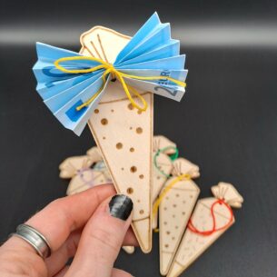 Eine Hand hält eine kleine Schultüte aus Pappelholz, an der ein blau gefalteter 20-Euro-Schein mit einer gelben Schnur befestigt ist. Im Hintergrund liegen weitere Holz-Schultüten mit verschiedenfarbigen Bändern