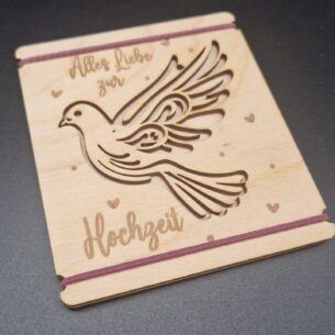 Holzkarte aus hellem Birkensperrholz mit dem lasergeschnittenen Motiv einer Taube und der Gravur „Alles Liebe zur Hochzeit“. Zwei lila Gummibänder halten die zwei Holzplatten zusammen. Die Karte liegt auf einem dunklen Untergrund.