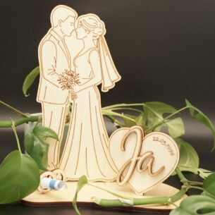 Lasergeschnittener Holz-Aufsteller aus hellem Birkensperrholz. Links ein küssendes Brautpaar in Hochzeitskleidung. Rechts daneben ein Herz mit dem 3D-Schriftzug „Ja“ und der gravierten Datumsangabe. Auf der Grundplatte steckt ein kleiner Holzring, in dem ein zusammengerollter Geldschein befestigt ist. Der Aufsteller steht vor einem dunklen Hintergrund mit grünen Blättern.