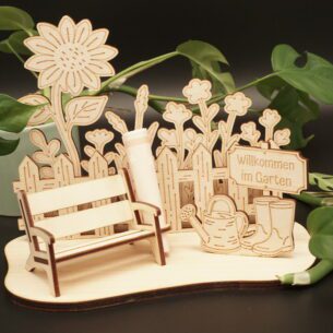 Detailansicht eines lasergeschnittenen Holz-Aufstellers, der eine Gartenszene darstellt. Links steht eine kleine Gartenbank mit einer Halterung für einen gerollten Geldschein dahinter. Im Hintergrund ist ein Lattenzaun mit Blumen und einer großen Sonnenblume zu sehen. Rechts stehen eine Gießkanne und Gummistiefel sowie ein Holzschild mit der Gravur „Willkommen im Garten“. Der Aufsteller ist aus hellem Birkensperrholz gefertigt und steht auf einer geschwungenen Grundplatte vor einem dunklen Hintergrund mit grünen Pflanzenblättern.