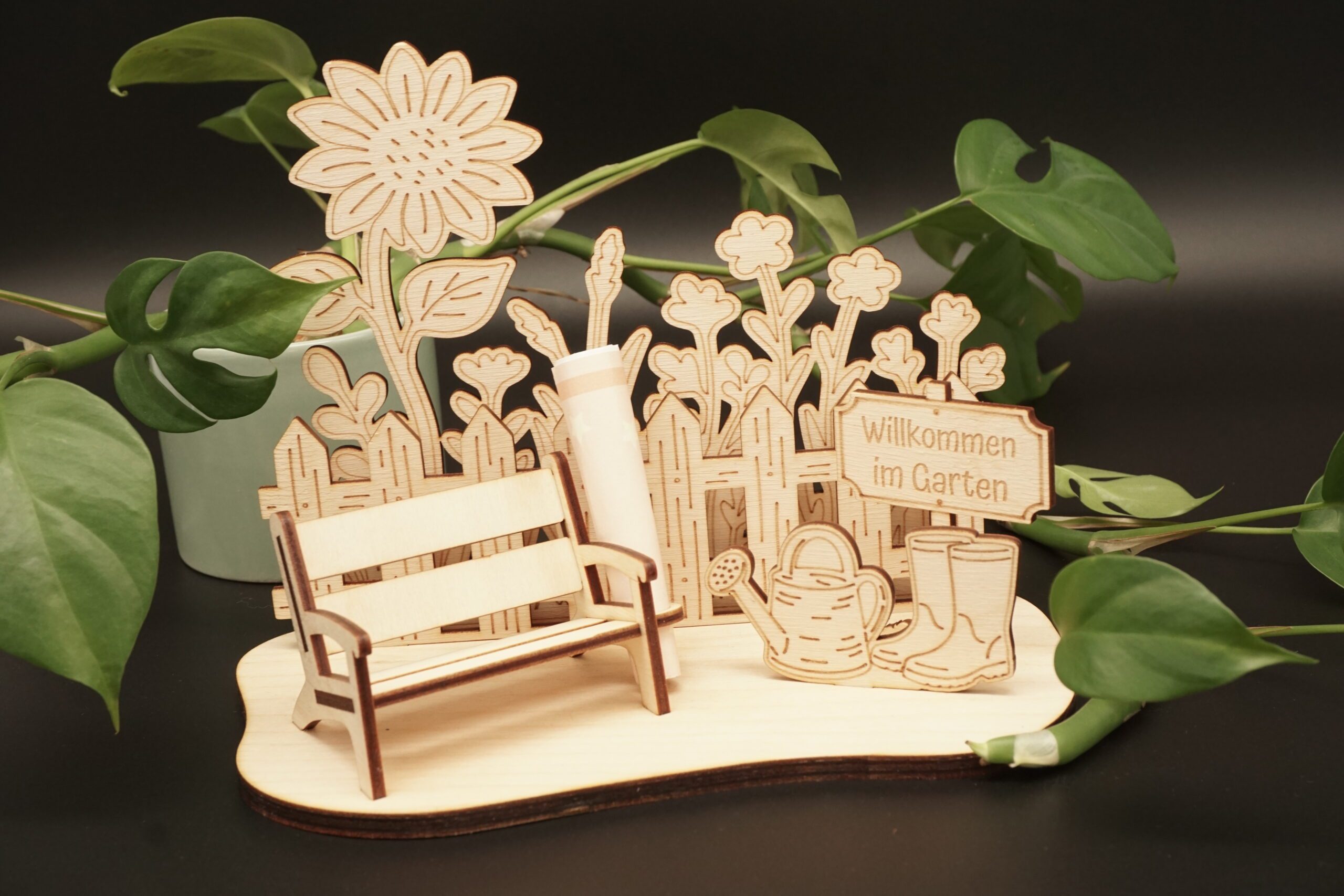 Detailansicht eines lasergeschnittenen Holz-Aufstellers, der eine Gartenszene darstellt. Links steht eine kleine Gartenbank mit einer Halterung für einen gerollten Geldschein dahinter. Im Hintergrund ist ein Lattenzaun mit Blumen und einer großen Sonnenblume zu sehen. Rechts stehen eine Gießkanne und Gummistiefel sowie ein Holzschild mit der Gravur „Willkommen im Garten“. Der Aufsteller ist aus hellem Birkensperrholz gefertigt und steht auf einer geschwungenen Grundplatte vor einem dunklen Hintergrund mit grünen Pflanzenblättern.