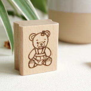 Draufsicht auf einen rechteckigen Holzstempel aus Buchenholz mit einer feinen Gravur eines sitzenden Bären-Mädchens, platziert auf hellem Untergrund neben grünen Blättern.