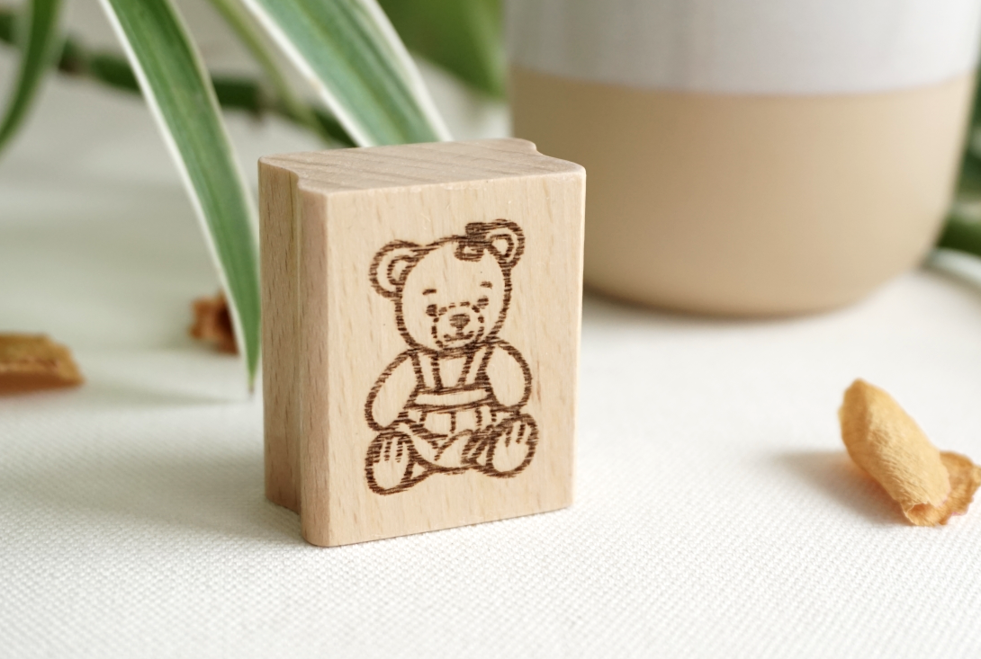 Draufsicht auf einen rechteckigen Holzstempel aus Buchenholz mit einer feinen Gravur eines sitzenden Bären-Mädchens, platziert auf hellem Untergrund neben grünen Blättern.