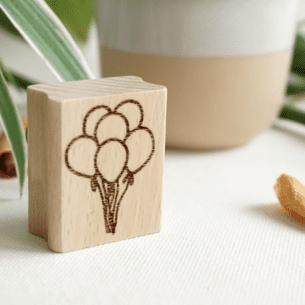 Senkrecht stehender Holzstempel aus Buchenholz mit einer Gravur von fünf zusammengebundenen Luftballons, im Hintergrund Dekoelemente und Pflanzen.
