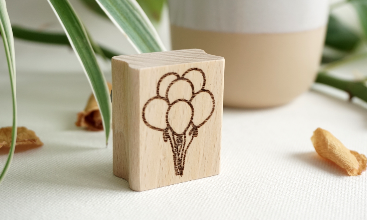 Senkrecht stehender Holzstempel aus Buchenholz mit einer Gravur von fünf zusammengebundenen Luftballons, im Hintergrund Dekoelemente und Pflanzen.