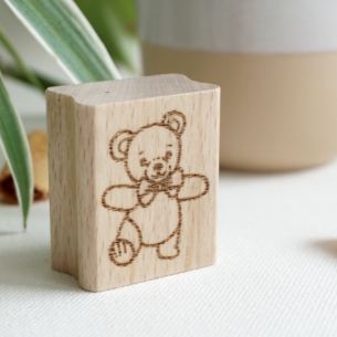 Hochkant stehender Holzstempel aus Buchenholz mit einer Gravur eines tanzenden, glücklichen Bärchens mit Schleife am Hals, im Hintergrund eine Pflanze und ein Keramikbecher.