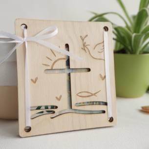 Quadratisches Geldgeschenk aus Buchenholz mit Kreuz-Ausschnitt und Gravuren von Fisch, Taube und Sonne, zusammengehalten durch eine weiße Schleife.