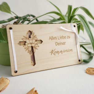Geldgeschenk zur Kommunion aus hellem Buchenholz mit ausgestanztem Kreuz, floraler Gravur und weißer Schleife vor grünem Pflanzenhintergrund.